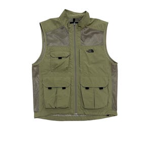 Può includere: Gilet verde oliva con cerniera a tutta lunghezza, tasche frontali multiple e pannelli laterali in rete. Il logo The North Face è sulla parte superiore sinistra del petto. Il gilet è senza maniche e ha un colletto alto.