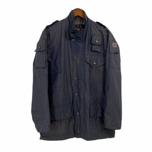 May include: A dark navy blue utility jacket featuring a high collar, a front button placket, and multiple flap pockets on the chest and sleeves. The jacket includes shoulder epaulets and a small rectangular patch on the left sleeve with a flag design.