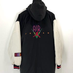 Pu&ograve; includere: Giacca da baseball nera e bianca con un grande logo "AIR JORDAN" sul retro. La giacca ha un cappuccio e maniche bianche con un logo "DREAM TEAM" colorato sulla manica sinistra.