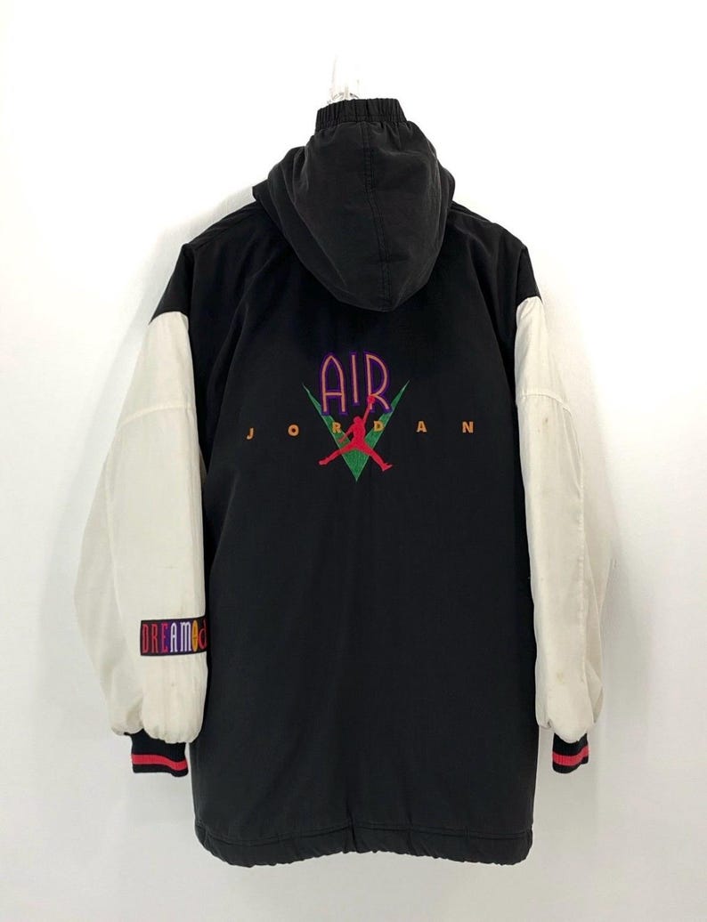 Pu&ograve; includere: Giacca da baseball nera e bianca con un grande logo "AIR JORDAN" sul retro. La giacca ha un cappuccio e maniche bianche con un logo "DREAM TEAM" colorato sulla manica sinistra.