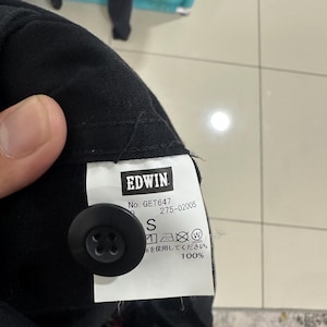 Pode incluir: Close-up de uma pe&ccedil;a de roupa preta com um bot&atilde;o preto e uma etiqueta branca. A etiqueta apresenta a marca "EDWIN", informa&ccedil;&otilde;es do produto e o tamanho "S". O tecido &eacute; 100% algod&atilde;o.
