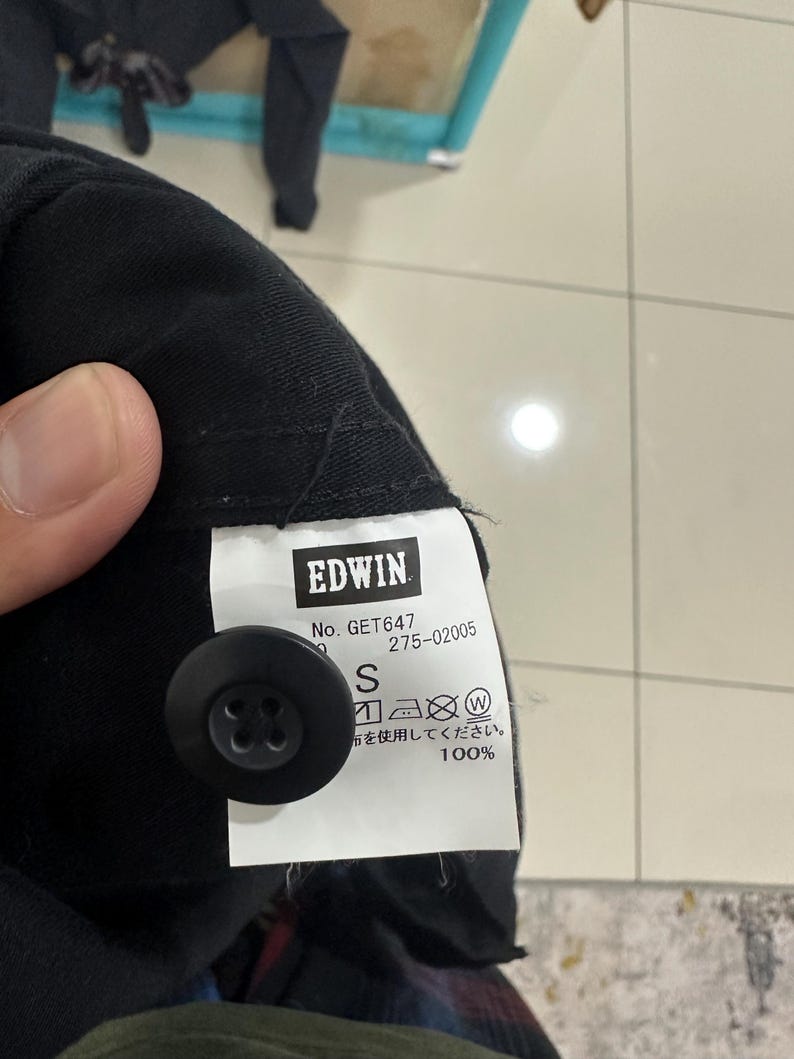 Pode incluir: Close-up de uma pe&ccedil;a de roupa preta com um bot&atilde;o preto e uma etiqueta branca. A etiqueta apresenta a marca "EDWIN", informa&ccedil;&otilde;es do produto e o tamanho "S". O tecido &eacute; 100% algod&atilde;o.
