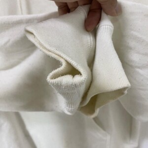 Puede incluir: Primer plano del tejido de algod&oacute;n blanco de la manga de una sudadera con capucha. El tejido est&aacute; doblado y muestra el detalle de la costura.