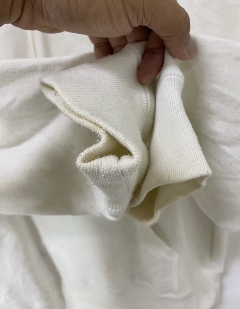 Puede incluir: Primer plano del tejido de algod&oacute;n blanco de la manga de una sudadera con capucha. El tejido est&aacute; doblado y muestra el detalle de la costura.