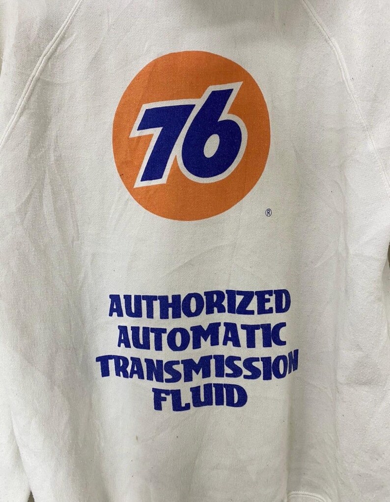 Puede incluir: Sudadera blanca con un gran c&iacute;rculo naranja que contiene el n&uacute;mero 76 en azul. Debajo del c&iacute;rculo est&aacute; el texto "AUTHORIZED AUTOMATIC TRANSMISSION FLUID" en azul.