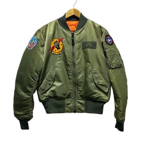 Peut inclure: Une veste bomber verte avec une fermeture éclair et plusieurs patchs. Les patchs comprennent un drapeau américain, un poing avec un éclair et un logo avec un croissant de lune.