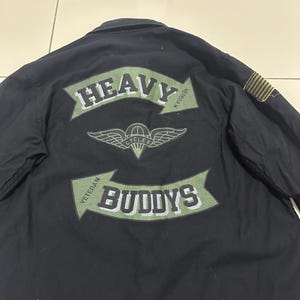 Pode incluir: Camisa preta com um gr&aacute;fico verde e branco nas costas que diz "HEAVY BUDDYS" com um emblema alado. A camisa tamb&eacute;m tem um pequeno patch na manga.