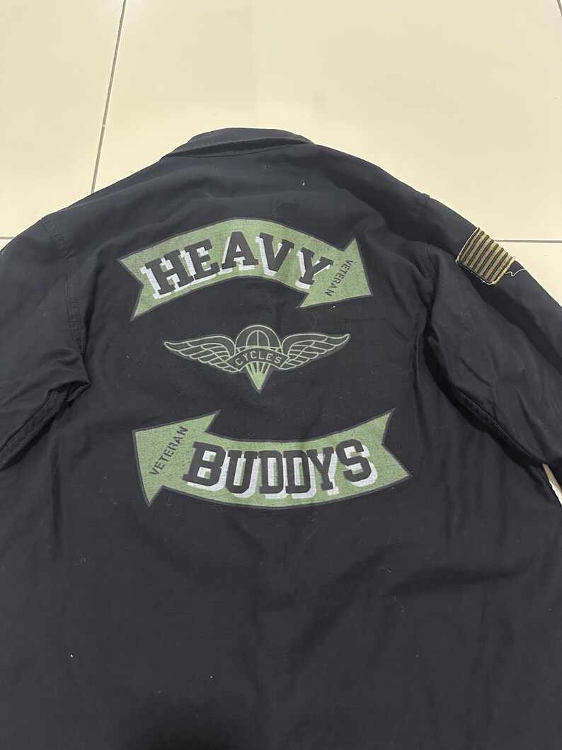 Pode incluir: Camisa preta com um gr&aacute;fico verde e branco nas costas que diz "HEAVY BUDDYS" com um emblema alado. A camisa tamb&eacute;m tem um pequeno patch na manga.
