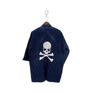 May include: Dark blue denim jacket with a white skull and crossbones graphic on the back. The jacket has a diamond pattern on the lower back and is hanging on a wooden hanger.
