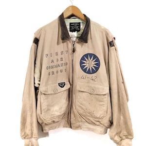 Puede incluir: Chaqueta bomber color beige con cuello marrón y múltiples bolsillos. La chaqueta presenta el texto "FIRST AIR COMMANDO GROUP" y un emblema azul y blanco. El número "41-42" también es visible. Cuelga de una percha de madera.