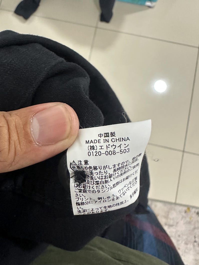 Pode incluir: Uma pe&ccedil;a de roupa preta com uma etiqueta branca com texto japon&ecirc;s e as palavras "MADE IN CHINA". A etiqueta tamb&eacute;m inclui um n&uacute;mero de telefone e instru&ccedil;&otilde;es de cuidados. A pe&ccedil;a de roupa &eacute; segurada por uma pessoa.