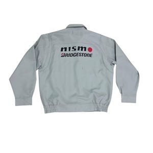 Puede incluir: Chaqueta gris claro con el texto "NISMO BRIDGESTONE" impreso en la espalda. La chaqueta tiene mangas largas, cuello y dobladillo acanalado. El logotipo de NISMO incluye un círculo rojo.