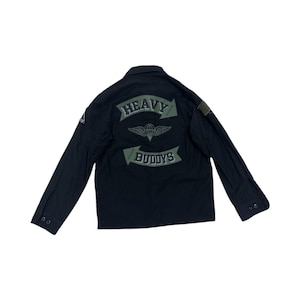 Pode incluir: Casaco preto de estilo militar com mangas compridas e fecho de bot&otilde;es. As costas apresentam um emblema verde com as palavras "HEAVY BUDDYS" e um desenho alado. Os patches s&atilde;o vis&iacute;veis nas mangas. O casaco est&aacute; estendido sobre uma superf&iacute;cie branca.