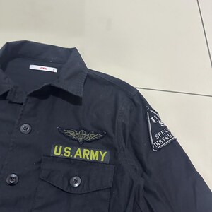 Pode incluir: Camisa preta abotoada com gola e dois bolsos no peito. A camisa apresenta um patch que diz "U.S. ARMY" em letras amarelas, juntamente com um emblema alado e um patch "U.S. SPECIAL INSTRUCTION". A camisa tem bot&otilde;es pretos.