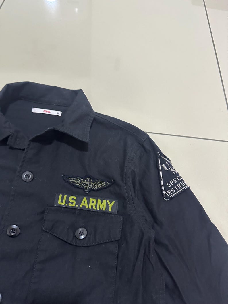 Pode incluir: Camisa preta abotoada com gola e dois bolsos no peito. A camisa apresenta um patch que diz "U.S. ARMY" em letras amarelas, juntamente com um emblema alado e um patch "U.S. SPECIAL INSTRUCTION". A camisa tem bot&otilde;es pretos.