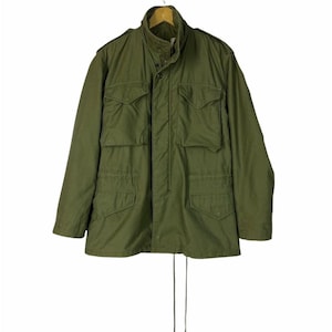 May include: An olive green military-style jacket with multiple front pockets and a drawstring waist. The jacket has a button-up front and a high collar. It is hanging on a wooden hanger against a white background.