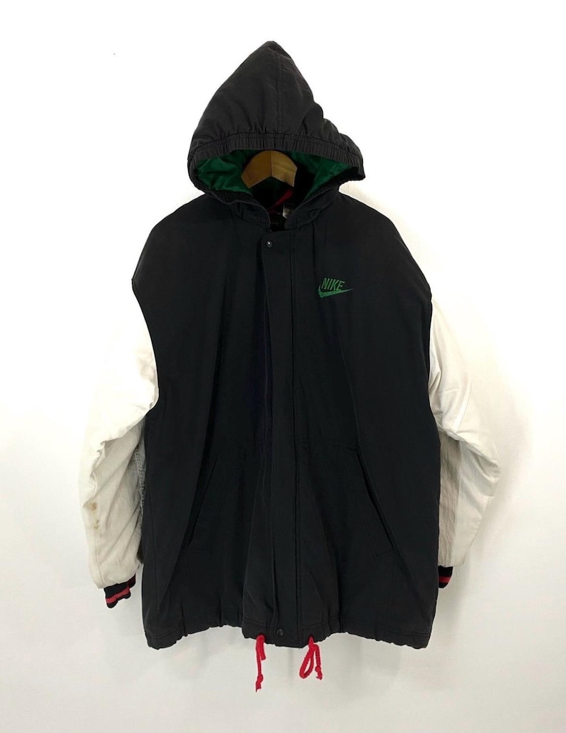 Pu&ograve; includere: Giacca con cappuccio nera e bianca con un logo Nike verde sul davanti. La giacca ha una manica bianca e un cordoncino rosso.