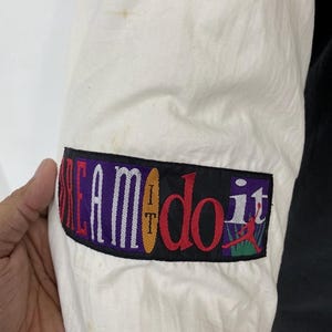 Pu&ograve; includere: Giacca bianca e nera con una toppa colorata sulla manica che dice "I Dream It Do It" con una grafica di un giocatore di basket.