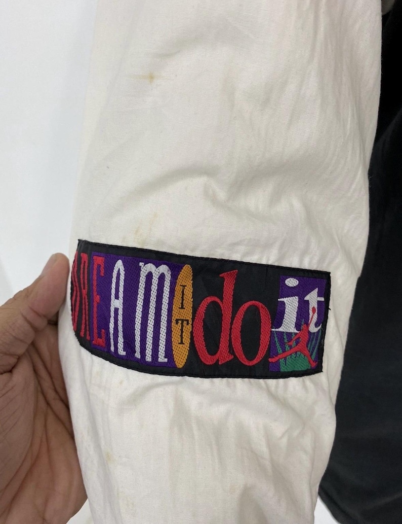 Pu&ograve; includere: Giacca bianca e nera con una toppa colorata sulla manica che dice "I Dream It Do It" con una grafica di un giocatore di basket.