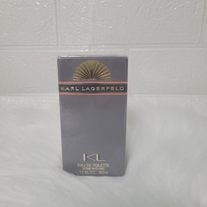 May include: A grey and silver perfume bottle with a pink and gold sunburst design. The bottle is labelled "Karl Lagerfeld" and "IKL Eau de Toilette Spray Naturel 1.7 fl oz 50 ml".