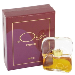 May include: A small, clear glass bottle of perfume with a gold cap. The bottle is shaped like a heart and has the word "Osé" printed on it. The bottle is sitting on a burgundy box with the words "J'ai Osé Parfum Paris" printed on it.