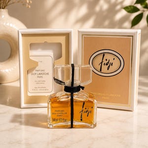 May include: A clear glass bottle of Fidji eau de toilette by Parfums Guy Laroche, containing 14 ml or 1/2 fl. oz. The bottle features a black cord tied around the neck and is presented with its original white and beige box packaging.