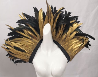 May include: A black and gold feather shoulder piece. The feathers are arranged in a fan shape and are attached to a black fabric strap.