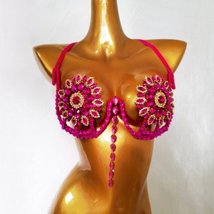 May include: A pink bra with a jeweled design. The bra has a pink velvet strap and a pink and gold jeweled design. The bra is on a gold mannequin.