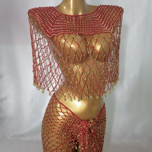 May include: A red and gold beaded two-piece outfit. The top is a net-like design with gold beads, and the skirt is a matching net-like design with gold beads. The outfit is displayed on a gold mannequin.