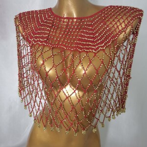 May include: A red and gold beaded shawl displayed on a mannequin. The shawl is made of red netting with gold beads. The beads are arranged in a diamond pattern. The shawl has a rounded neckline and hangs over the shoulders.