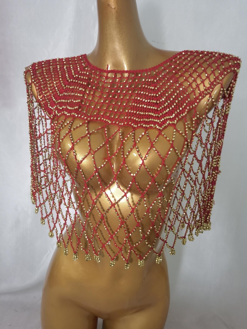 May include: A red and gold beaded shawl displayed on a mannequin. The shawl is made of red netting with gold beads. The beads are arranged in a diamond pattern. The shawl has a rounded neckline and hangs over the shoulders.