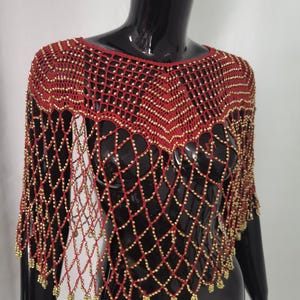 May include: A red crocheted shawl with gold-coloured beads. The openwork design features a diamond pattern, with the beads accenting the intersections. The shawl is displayed on a black mannequin.