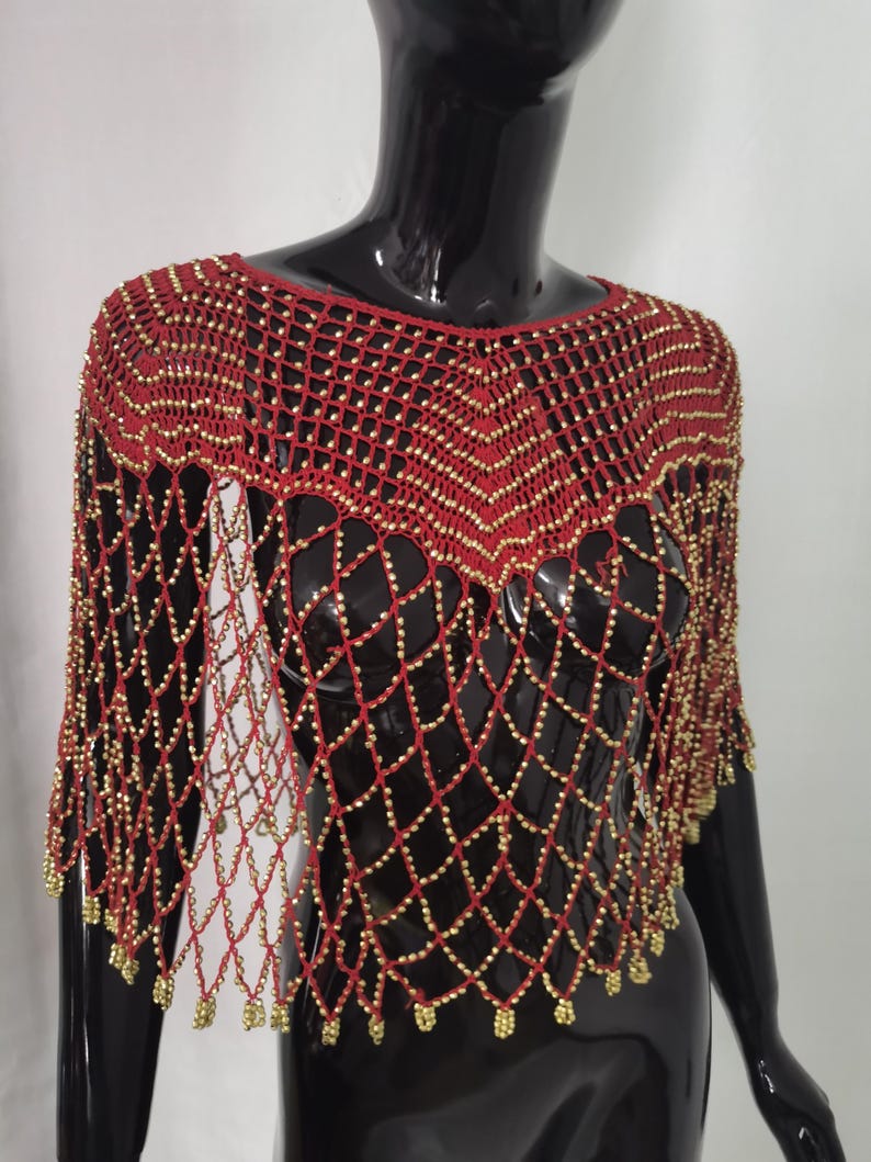 May include: A red crocheted shawl with gold-coloured beads. The openwork design features a diamond pattern, with the beads accenting the intersections. The shawl is displayed on a black mannequin.