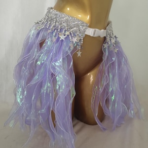 May include: A lavender and silver belly dance skirt with a white belt. The skirt features a silver beaded waistband with dangling star charms and flowing lavender fabric with an iridescent sheen. The skirt is designed for dance or costume.