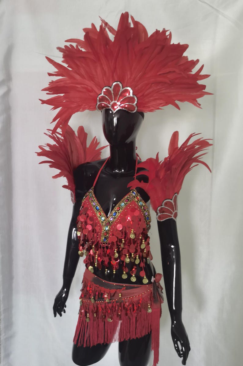 May include: A vibrant red carnival costume with a feathered headdress, shoulder pieces, and a sequined top and skirt. The costume features gold and jewel accents, and fringe detailing. The outfit is displayed on a black mannequin.