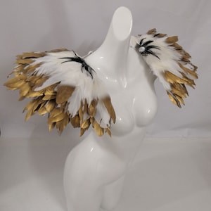 Puede incluir: Pieza de hombro de plumas blancas y doradas con detalles negros. Las plumas est&aacute;n dispuestas en un dise&ntilde;o en capas y en cascada.
