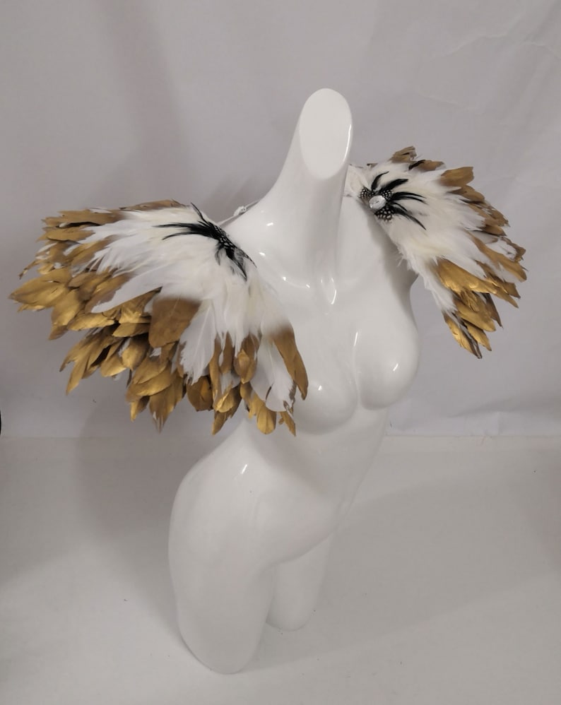 Puede incluir: Pieza de hombro de plumas blancas y doradas con detalles negros. Las plumas est&aacute;n dispuestas en un dise&ntilde;o en capas y en cascada.