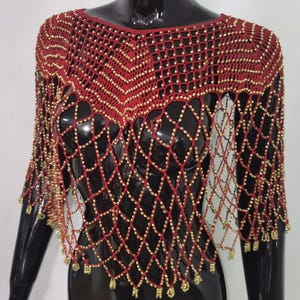May include: A red and gold beaded shawl with a net-like design. The shawl has a round neckline and short sleeves. Gold beads are evenly spaced throughout the red netting, creating a shimmering effect. The garment is displayed on a black mannequin.