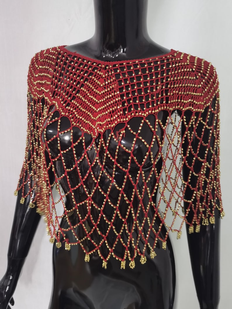 May include: A red and gold beaded shawl with a net-like design. The shawl has a round neckline and short sleeves. Gold beads are evenly spaced throughout the red netting, creating a shimmering effect. The garment is displayed on a black mannequin.