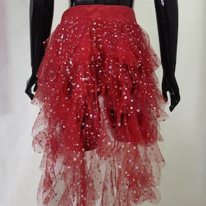 Puede incluir: Falda roja de tul con múltiples capas y lentejuelas dispersas. La falda tiene un dobladillo asimétrico, con la parte trasera más larga que la delantera. La falda se muestra en un maniquí negro.