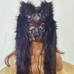 Op de afbeelding: Een zwart wolfsmasker met een gedetailleerd leren gezicht en een capuchon van imitatiebont. Het masker heeft een gestileerd wolfsgezicht met scherpe tanden en een rode lip. De capuchon loopt naar beneden met lang, zwart imitatiebont.