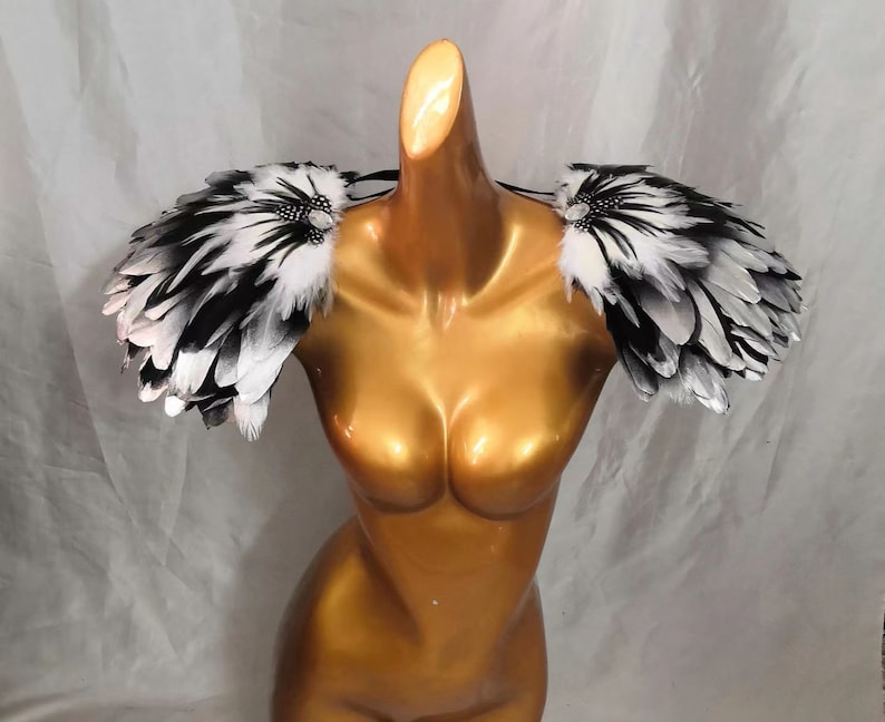 Puede incluir: Un par de hombreras de plumas negras y blancas con pedrer&iacute;a. Las hombreras se llevan sobre un maniqu&iacute; con un cuerpo de color dorado.