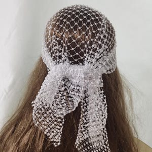 May include: A white bridal headpiece featuring a net design, covering the top of the head. The headpiece is secured with a white ribbon tied in a bow at the back. The net is dotted with small, white beads.