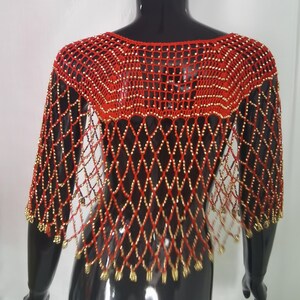 May include: A red and gold beaded shawl on a black mannequin. The shawl has a diamond-patterned net design with gold beads at the intersections and edges. The neckline is a scoop, and the sleeves are short. The shawl is a striking accessory.