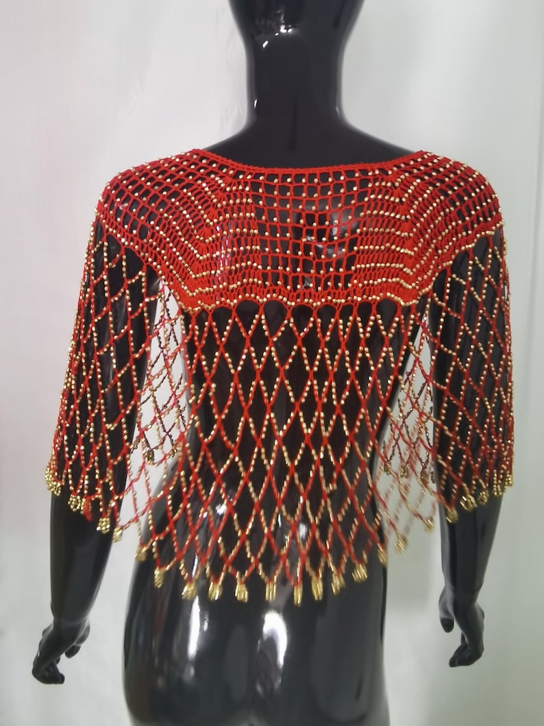May include: A red and gold beaded shawl on a black mannequin. The shawl has a diamond-patterned net design with gold beads at the intersections and edges. The neckline is a scoop, and the sleeves are short. The shawl is a striking accessory.