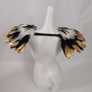 Puede incluir: Un par de hombreras de plumas negras y blancas con detalles dorados. Las hombreras est&aacute;n unidas a una correa negra.