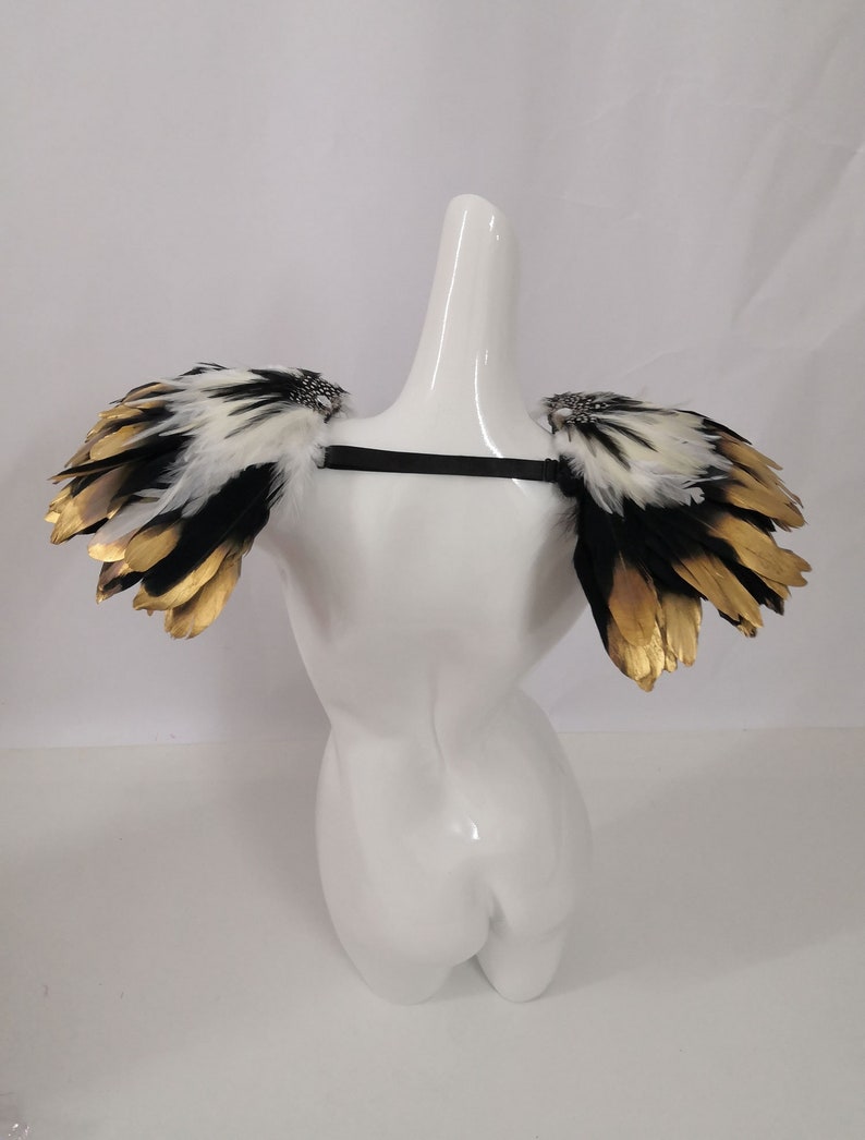 Puede incluir: Un par de hombreras de plumas negras y blancas con detalles dorados. Las hombreras est&aacute;n unidas a una correa negra.