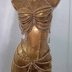 May include: A gold-toned body chain accessory featuring multiple draped strands of metallic beads. The design includes a halter-style top section with layered bead chains and a matching lower waist section, all connected by thin metallic chains.