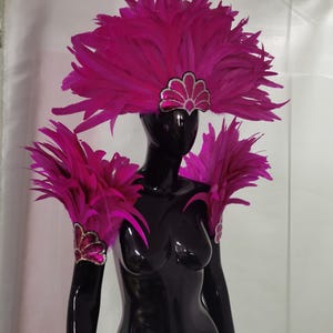 May include: A black mannequin displays a vibrant fuchsia feather headdress and shoulder pieces. The headdress features a silver and pink sequined accent. The shoulder pieces and arm cuffs are adorned with matching feathers and sequin details, creating a dramatic, eye-catching ensemble.