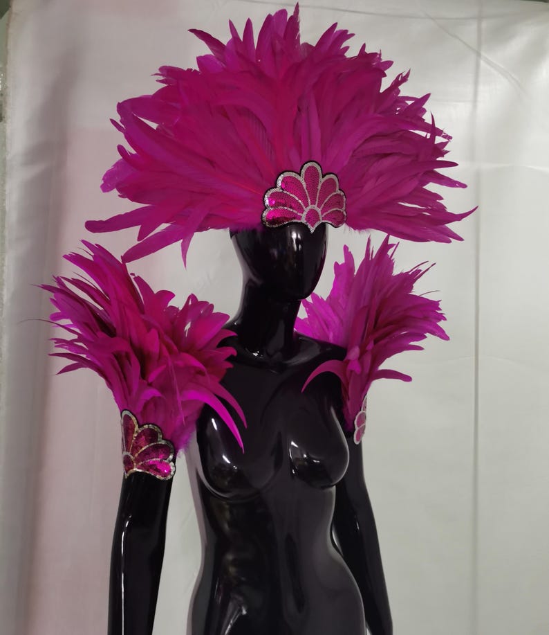 May include: A black mannequin displays a vibrant fuchsia feather headdress and shoulder pieces. The headdress features a silver and pink sequined accent. The shoulder pieces and arm cuffs are adorned with matching feathers and sequin details, creating a dramatic, eye-catching ensemble.